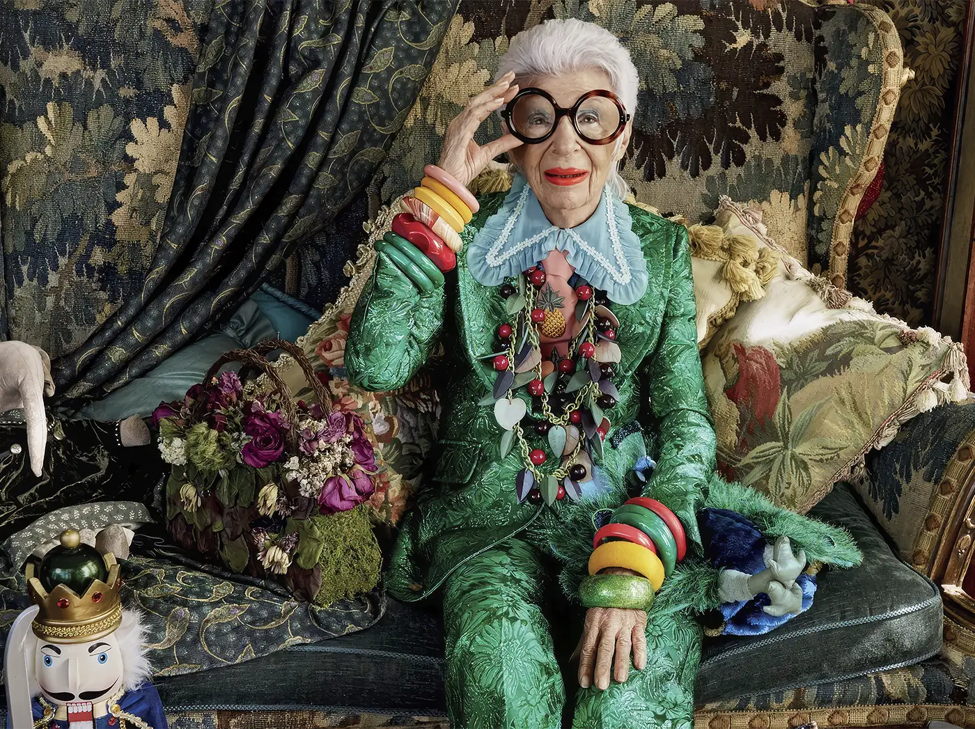 Iris Apfel wearing her iconic glasses, sitting on the sofa. Dressing Boldly, Living Boldly: Iris Apfel’s Treasures on Auction. US